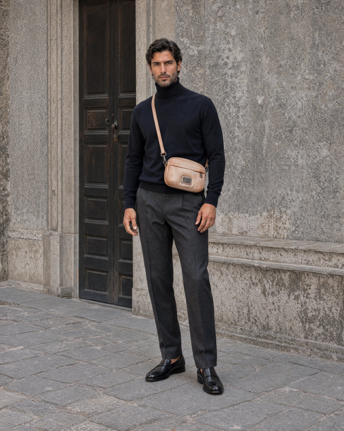 Man wearing a navy knit top with a tan distressed-leather crossbody bag in a stone courtyard