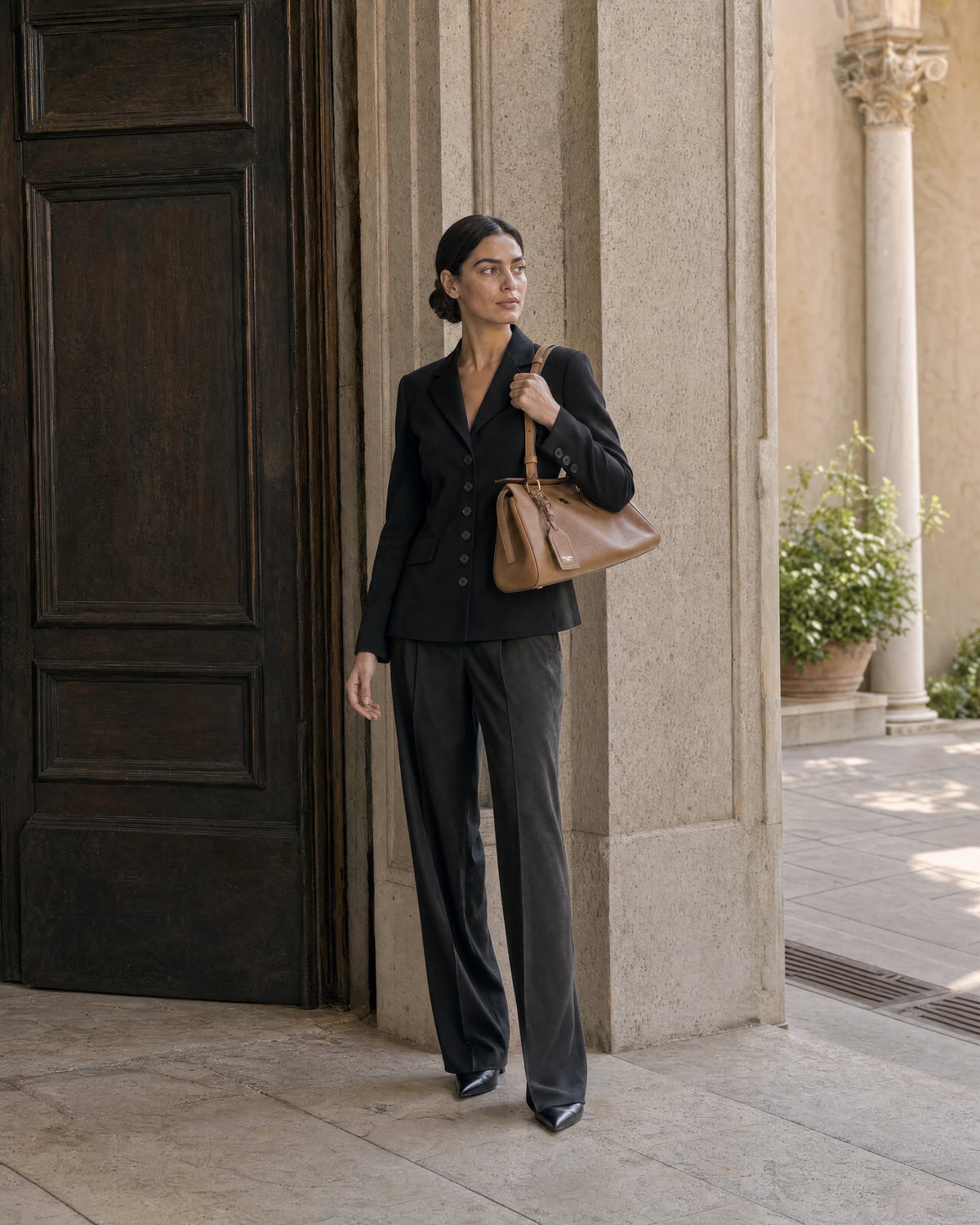Woman in black tailoring carrying a tan leather shoulder bag beside a stone doorway